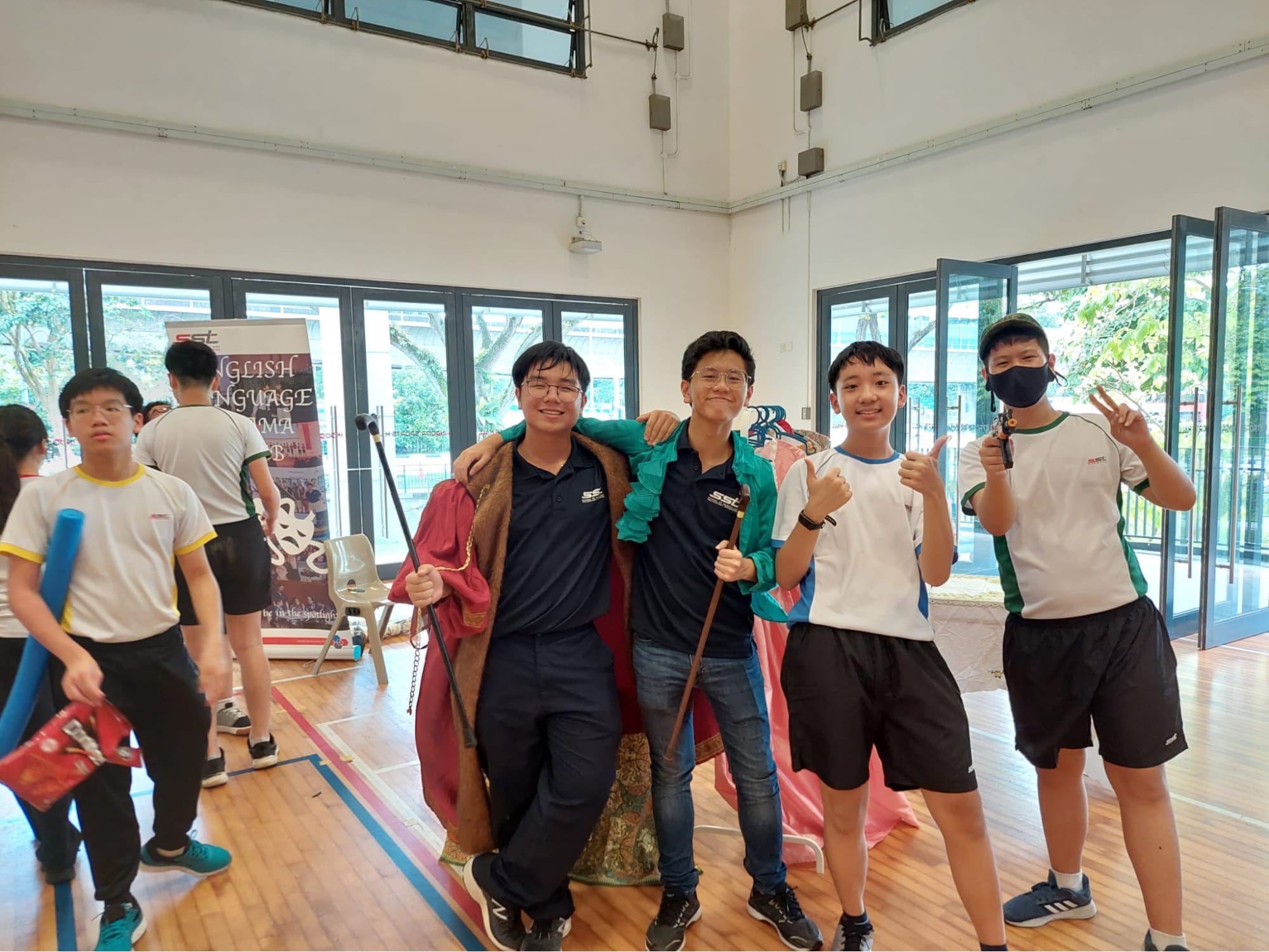 Four male students pose smiling in a school hall near an English Language Drama banner, some holding props and wearing costumes.